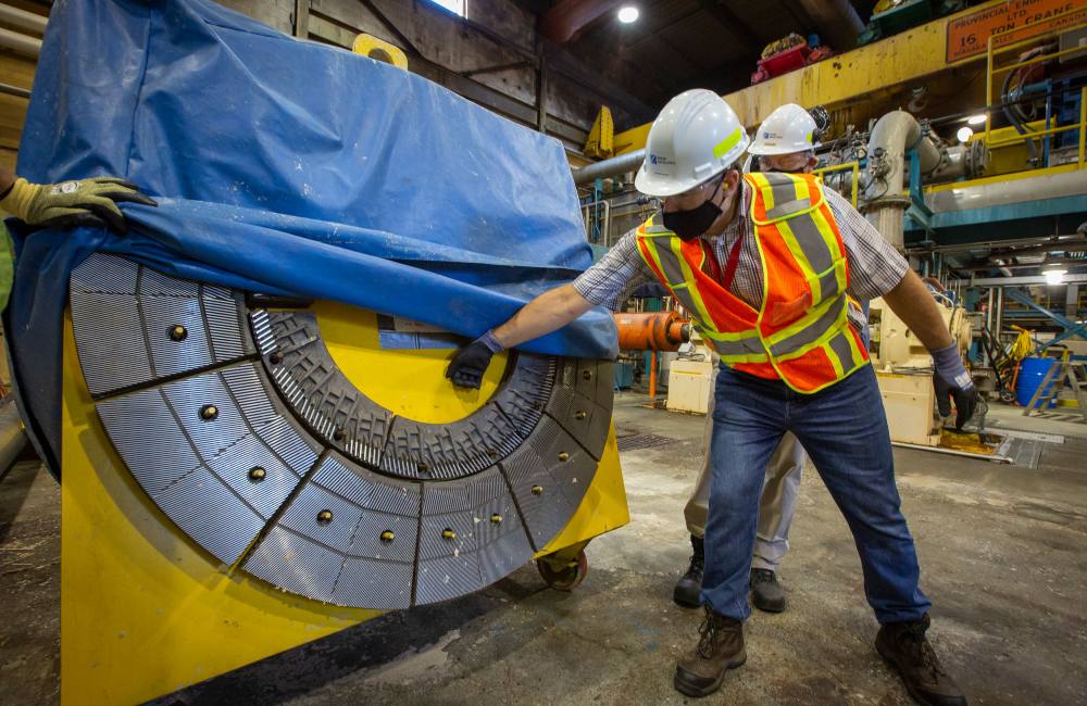 A man in a safety vest, a white hard hat and boots inspects a large mechanical machine part. The part is yellow with a metal gray circular component and a gloved hand holds up the blue tarp covering it.