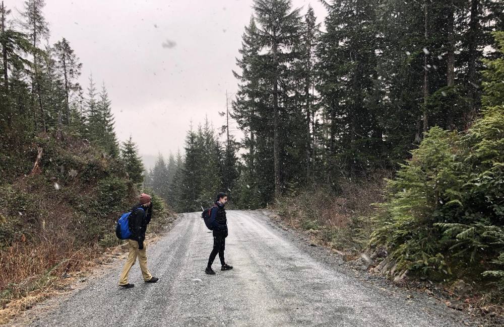 Two young men walk across a dirt road in the snow. The sky is grey and they are both wearing black rain jackets. The man on the left is wearing a brown toque and blue backpack. 