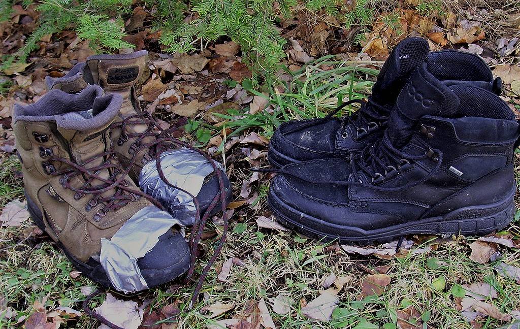 Two pairs of hiking boots on the ground with leaves and evergreen needles, one wrapped with silver duct tape.