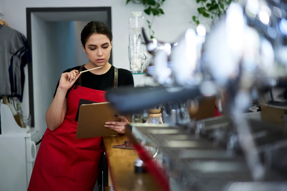 A dark haired young woman in an apron studies a clipboard behind the counter of a coffee shop.