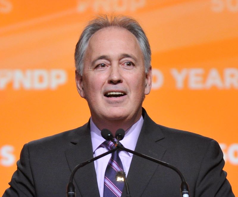 A man with thinning grey hair, wearing a dark suit, pale blue shirt and striped tie, stands at a podium in front of an orange sign that says BCNDP.