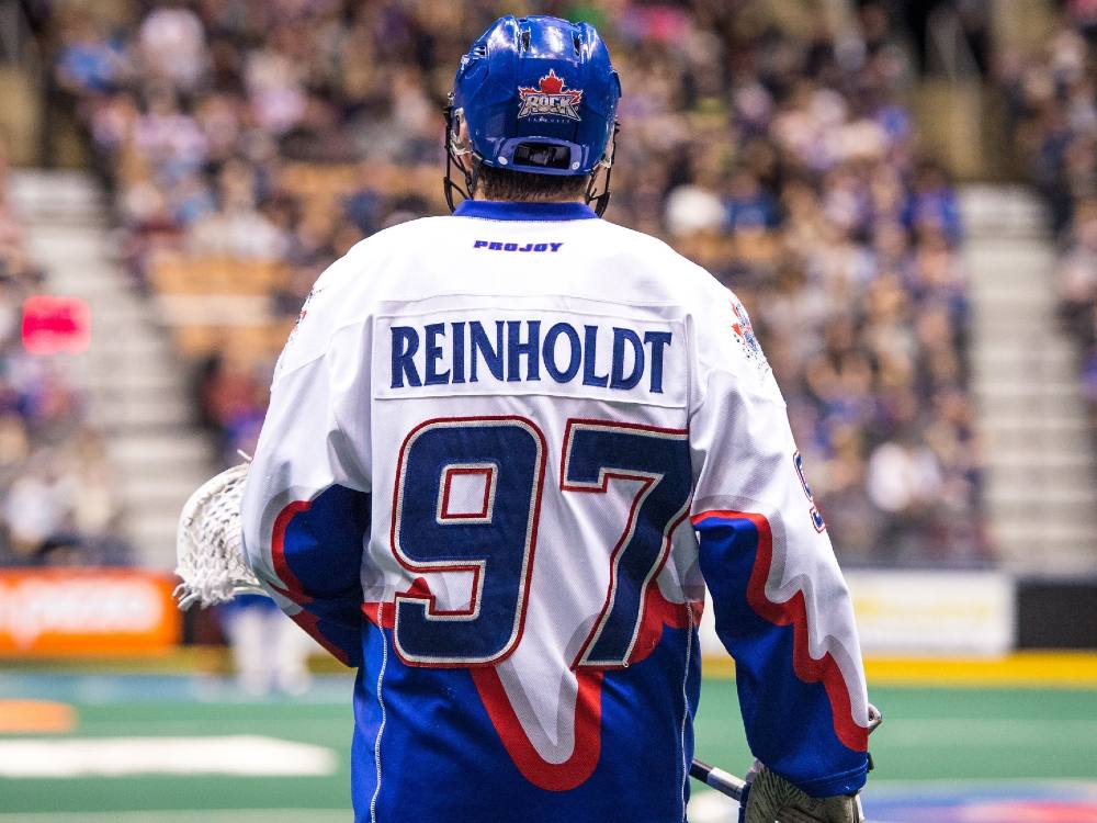A lacrosse player in full gear, red white and blue, seen from the back with the name Reinholdt across the shoulders.