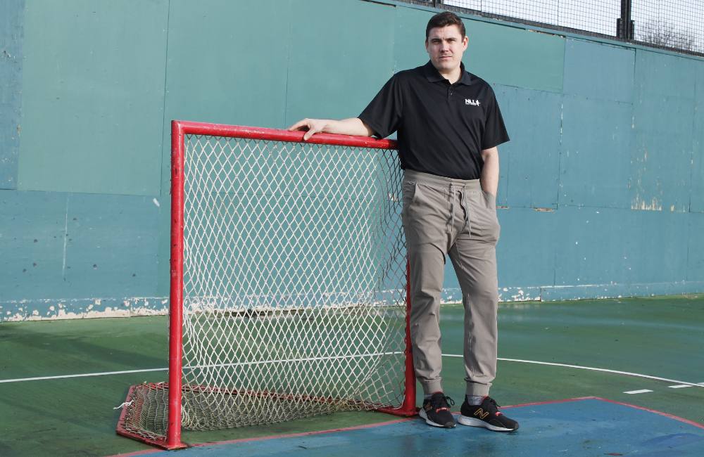 A man leans against a lacrosse net, wearing a black short-sleeved polo shirt and grey sweatpants.