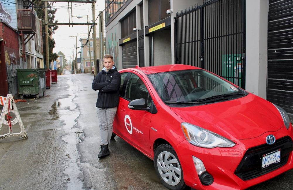 A man with short brown hair, wearing grey pants and a blck jacket, leans against a bright red Modo car in an alley.