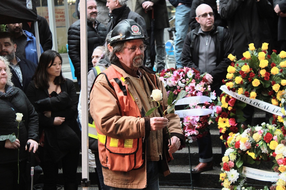 A man with longish hair and glasses stands in front of floral wreathes. He is wearing a black hardhat, a heavy coat and orange safety vest, and holds a single white rose.