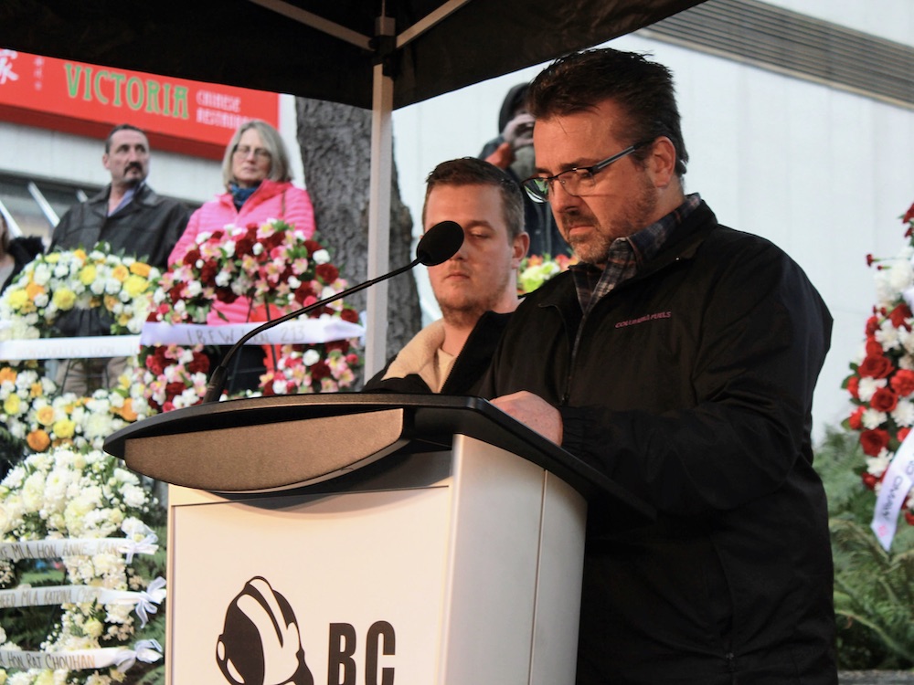 A man with short hair and glasses, wearing a dark jacket, sands at a podium. Large floral wreaths are on each side.