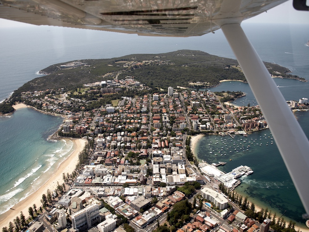 An aerial photo shows a peninsula, ringed with beaches, where a residential area transitions into bushland.