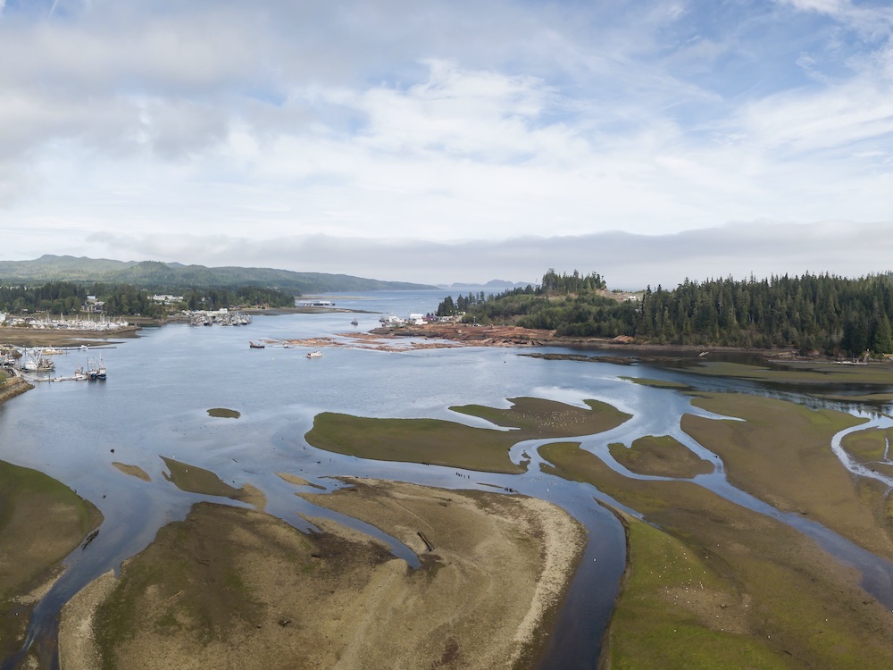 An aerial view of a small municipality on northern Vancouver Island depicts estuaries that open out onto the Pacific Ocean. Sprawling sand spits are in the foreground, and they converge into open water against a coastal mountain range in the background. The sky is blue with clouds.