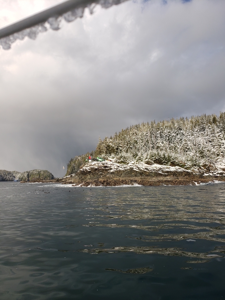 A photograph of a snowy, hilly island depicts a green helicopter landing at a blinker on a hill. The shot is taken from a boat on water. An icy railing is high in the foreground.