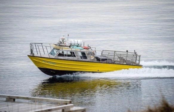A yellow boat with a grey metal cabin drives towards the left of the frame. Part of a dock is visible in the foreground. The water is grey.