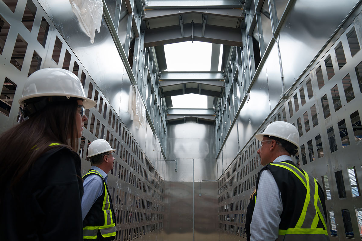 Three people wearing white hardhats stand in a towering room. The interior is clad in stainless steel. 