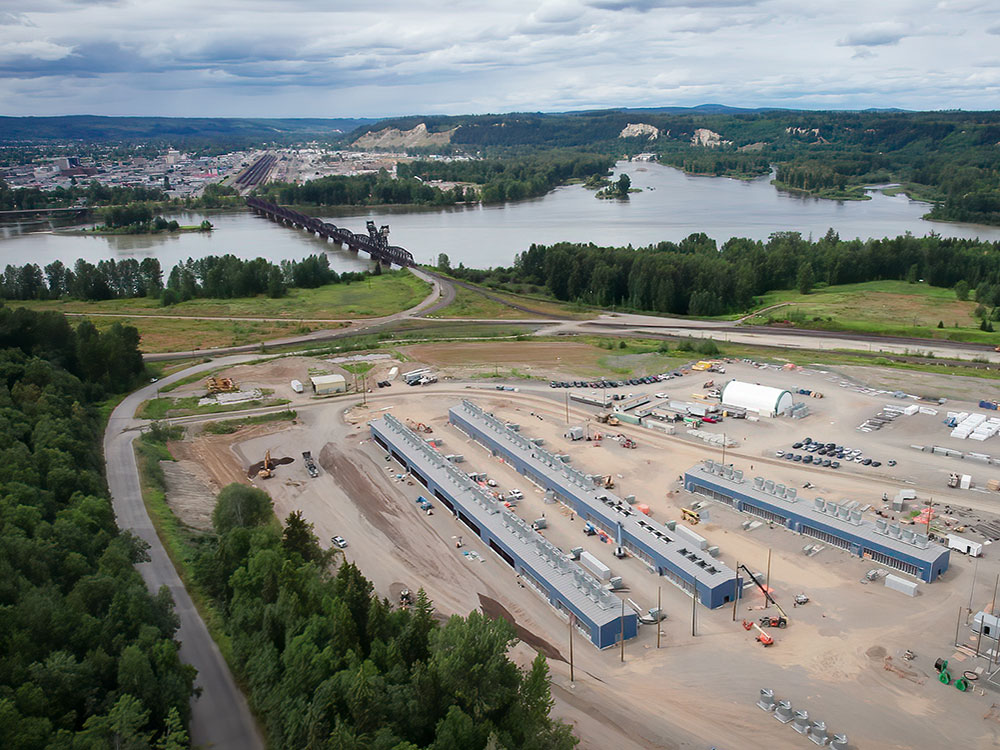 In the foreground, several long warehouses sit on an industrial site. Beyond there is a water body and an urban area surrounded by forest. 