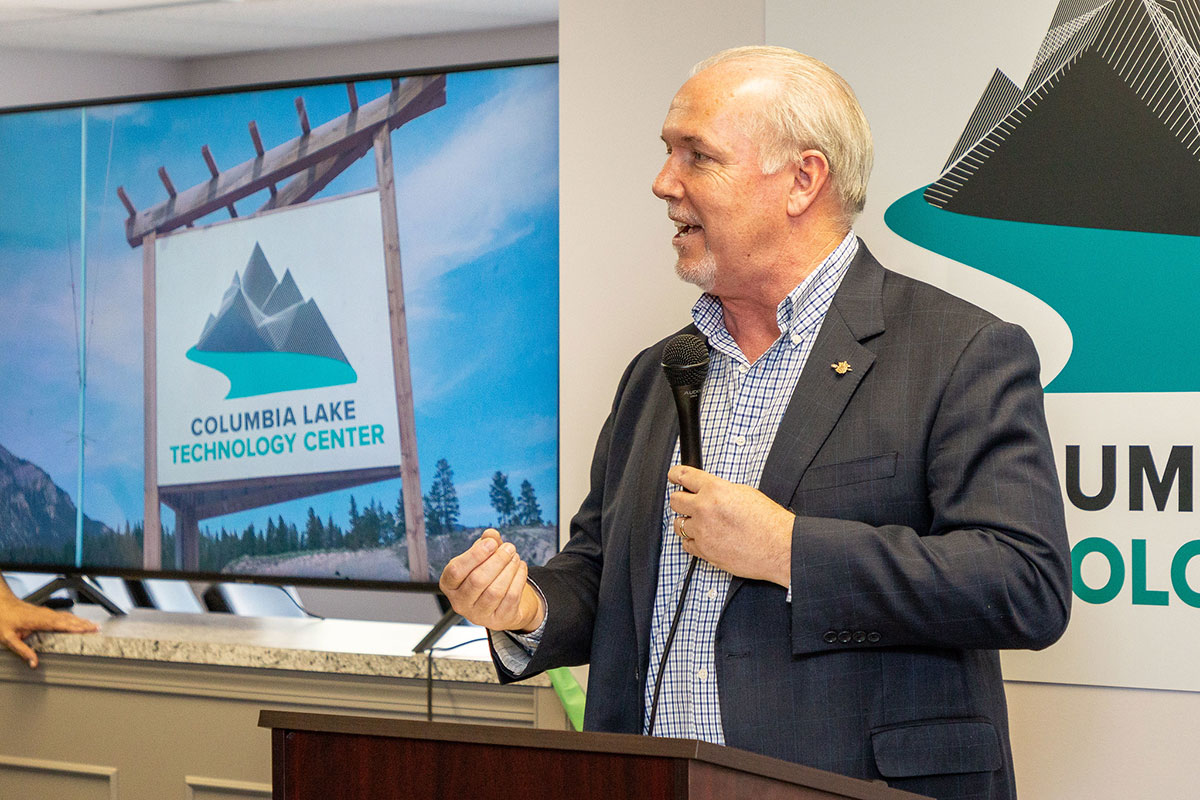 A balding man stands at a podium holding a microphone in one hand and gesturing with the other. Behind him, a large image shows a sign that reads Columbia Lake Technology Center.