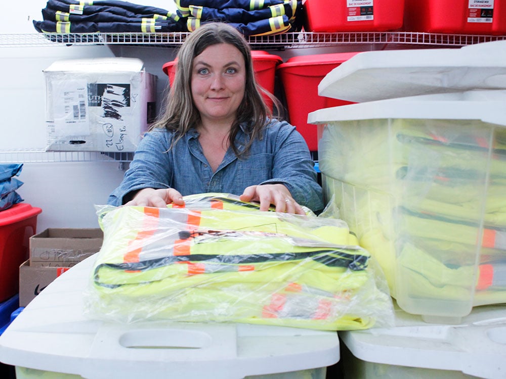 A woman with long brown hair wearing a blue shirt smiles at the camera, surrounded by bins and shelves holding safety vests and coveralls.