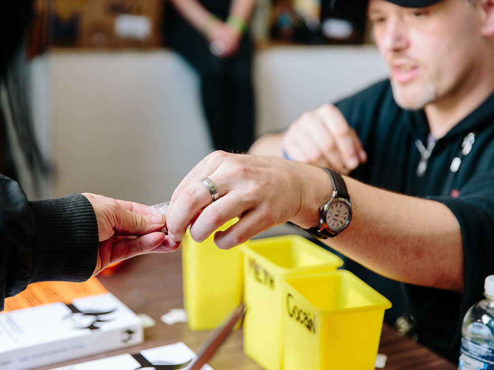 A man wearing a black shirt sits at a table with three yellow containers in front of him, one labelled cocaine and one labelled meth. He is passing a small plastic bag to another person’s outstretched hand.