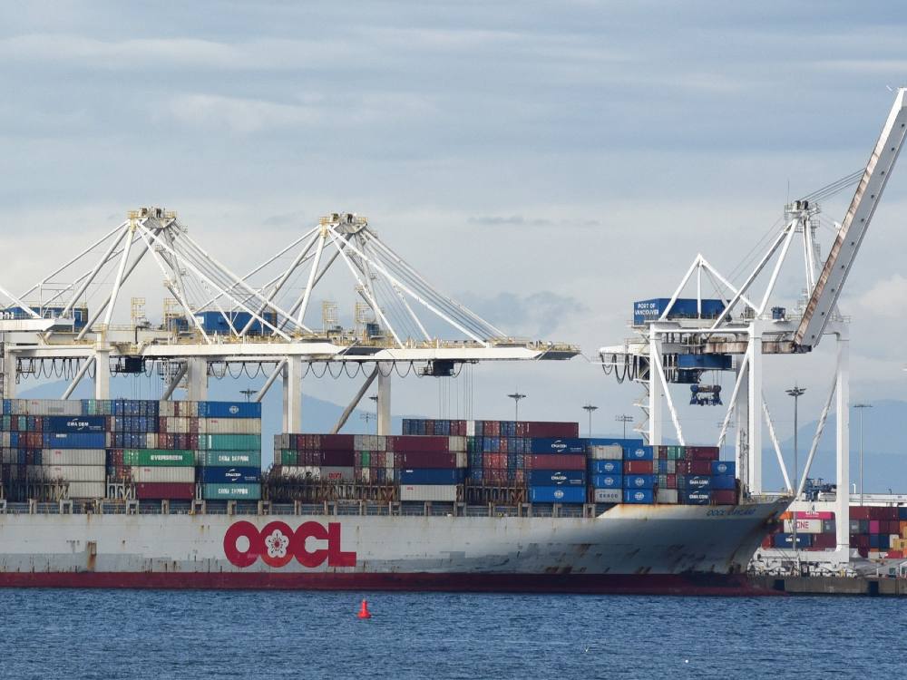 A large grey ship stacked high with containers is in the foreground. In the background giant white cranes loom over it.