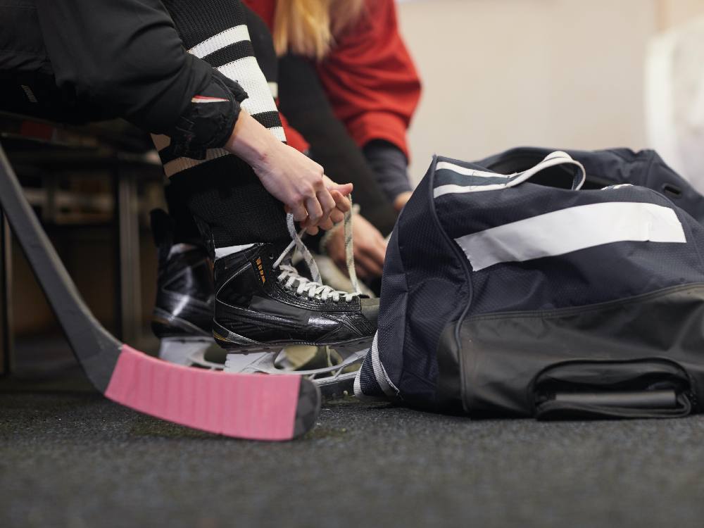 Kids lace up their skates in a hockey dressing room. A hockey stick is visible in the foreground, wrapped with pink tape. 