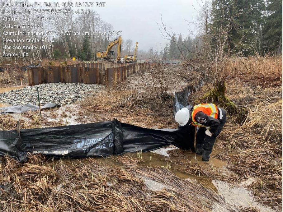  A person bends over a black fence made out of a plastic tarp material. They appear to be lifting up part of the fence that is allowing water to seep out of the construction area. Construction machinery is visible in the background.
