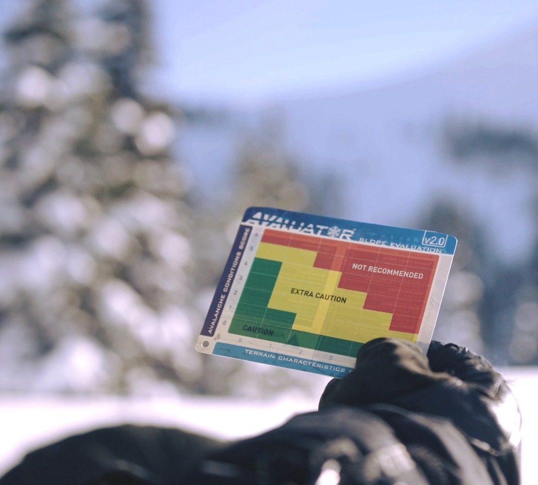 A gloved hand holds a card with red, yellow and green areas denoting avalanche risk. Blue sky and snowy trees are blurred in the background.