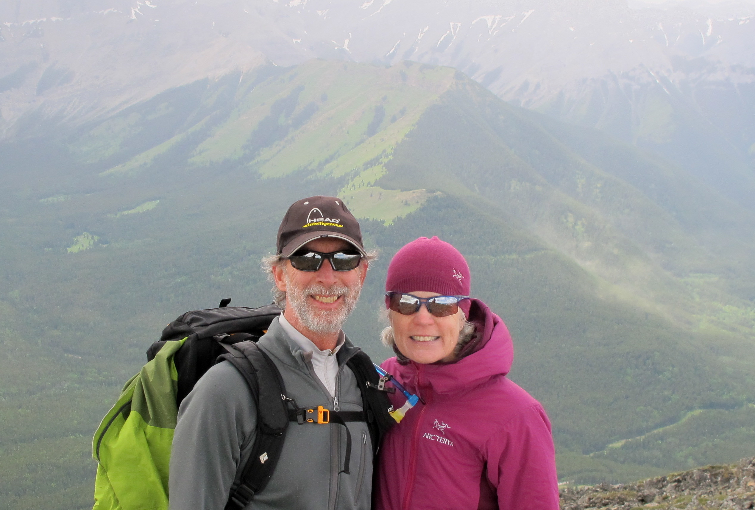 A man and a woman stand in a mountain setting. The man wears black ballcap, backpack and grey fleece hiking shirt. The woman wearing a matching pink tuque and athletic jacket. 