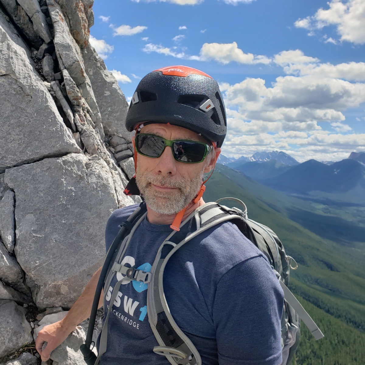 A man wearing a helmet and backpack stands next to a rock face high above a mountain valley.