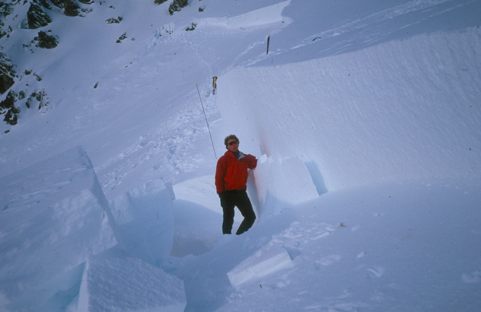A man in a red ski jacket stands in a mountain environment next to a vertical wall of snow that is almost twice his height.