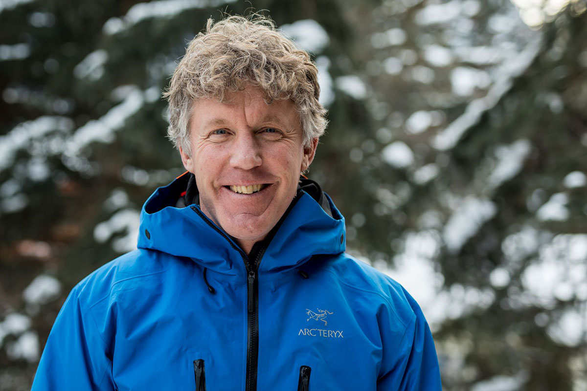 Portrait of a smiling man with greying hair wearing a blue ski jacket. He stands in front of forested background. 