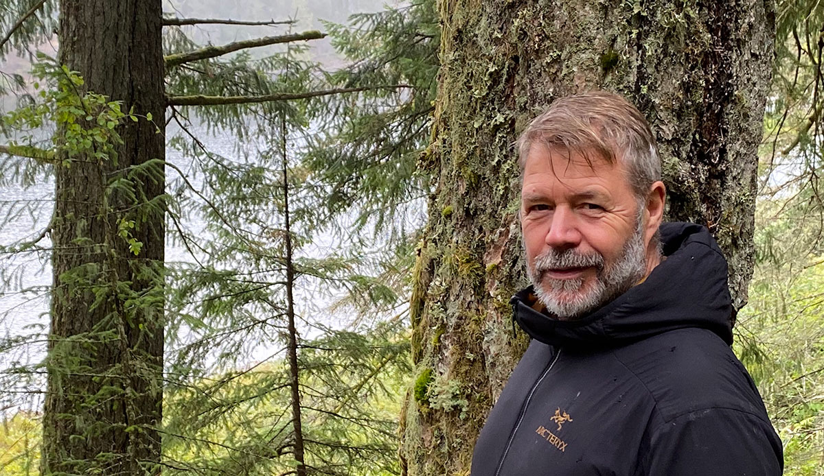 A portrait of a man with greying hair and beard wearing a black ski jacket standing in the forest with the ocean beyond.