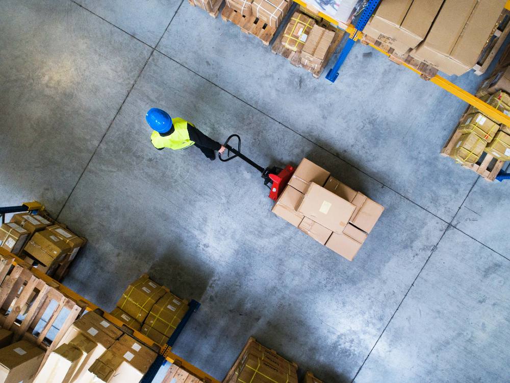 An overhead photo shows a worker wearing yellow safety vest and blue helmet pulling a load of cardboard boxes through a warehouse. Tall shelves rise on each side.