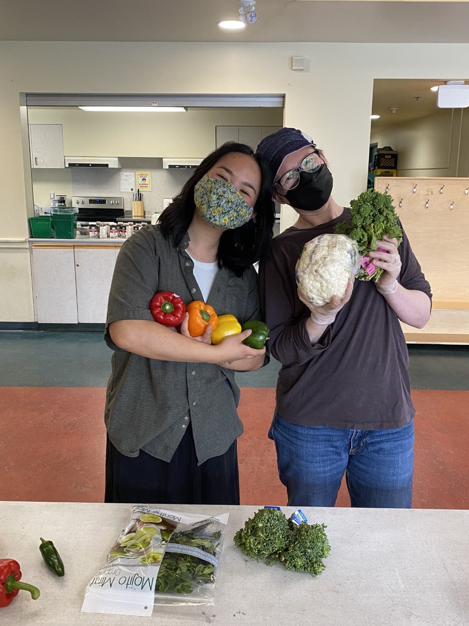 Two people holding produce (bell peppers, cauliflower and broccoli) and wearing masks smile and pose for a photo.