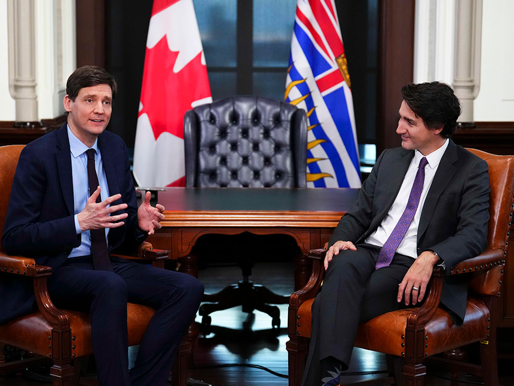 David Eby and Justin Trudeau sit in leather chairs in front of a desk and Canadian and B.C. flags. Both wear suits and ties. Eby is gesturing and Trudeau is smiling.