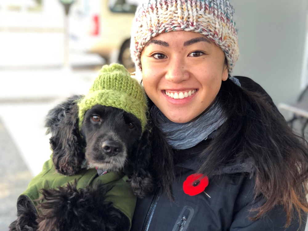 A woman wearing a toque and winter coat looks at the camera, at ease and smiling. Her dog, also wearing a toque and coat, sits on her lap. 