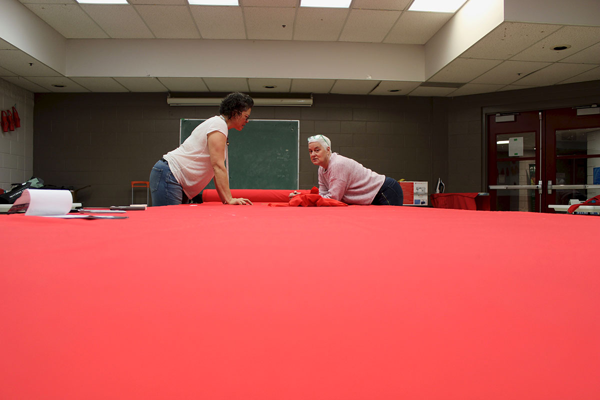 A long swath of red fabric covers a table. At the end of the table, two women lean over the fabric.