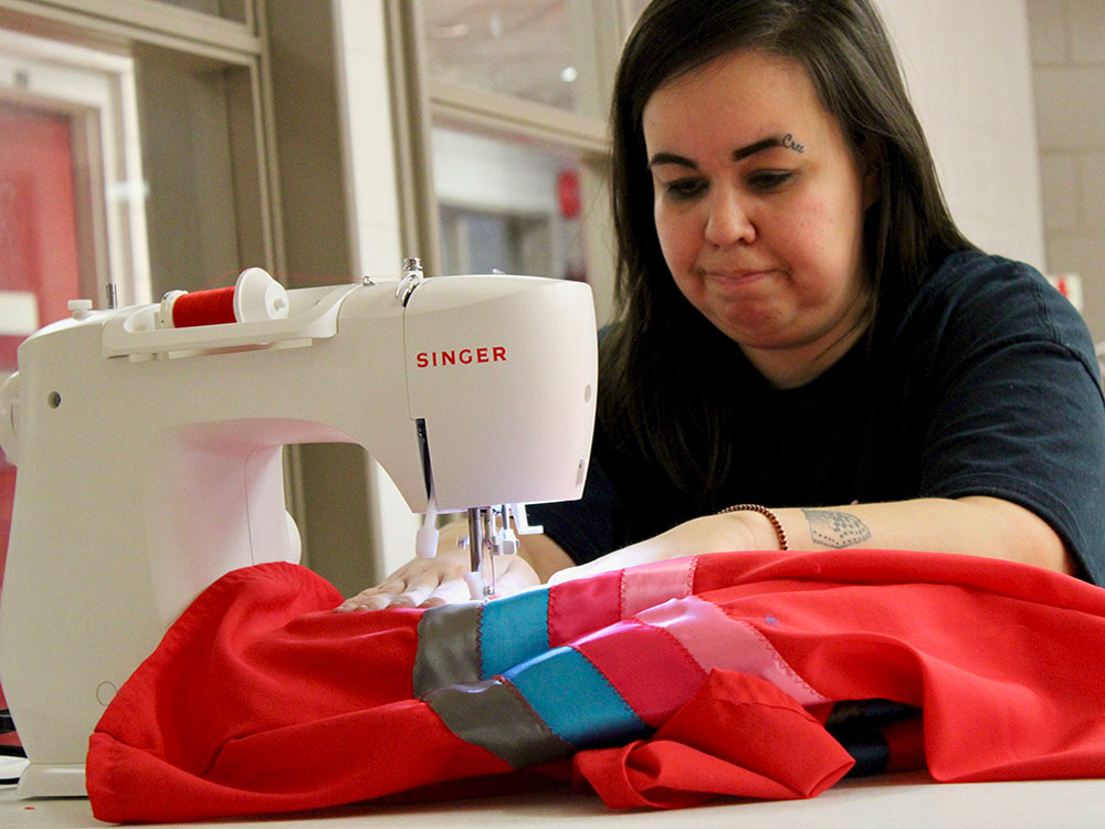 A woman sews red fabric and ribbons at a sewing machine. Her face shows an expression of concentration and she has a tattoo that reads &ldquo;Cree&rdquo; near her left eyebrow.