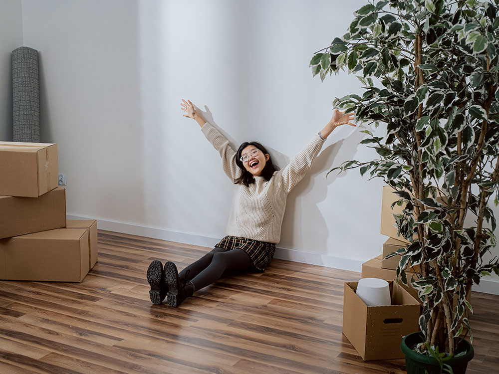 A young woman sits on the ground in a new apartment, surrounded by a few moving boxes. She raises her arms towards the sky.