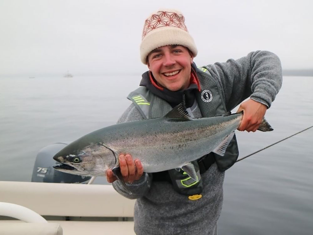 A smiling young man holds a large salmon. He is wearing a toque, sweater and life vest and is in a boat on calm sea.