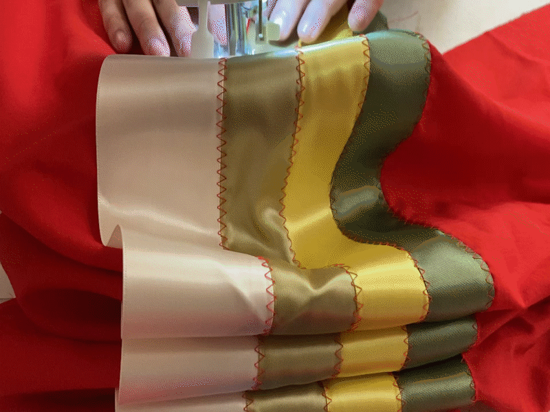 A woman&rsquo;s hands guide red fabric with ribbons sewn to it through a sewing machine.