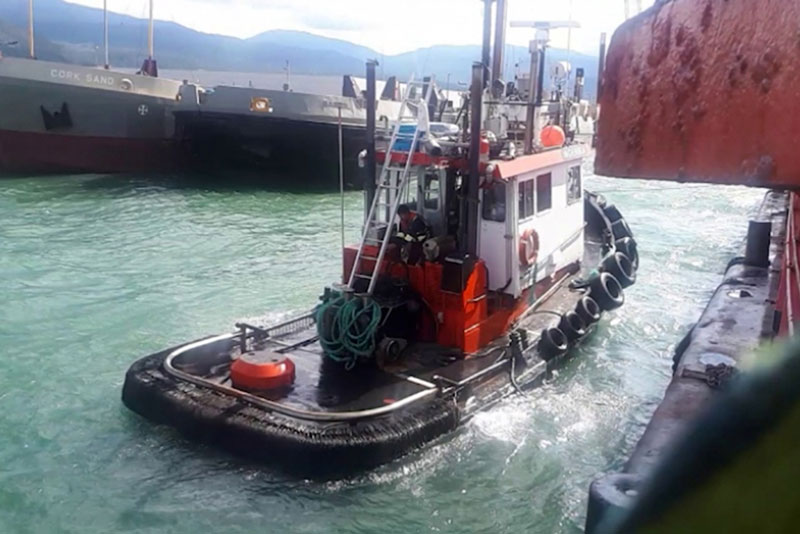 A tugboat with a black hull and red and white cabin sits near a dock. Old tires a fastened to the front.