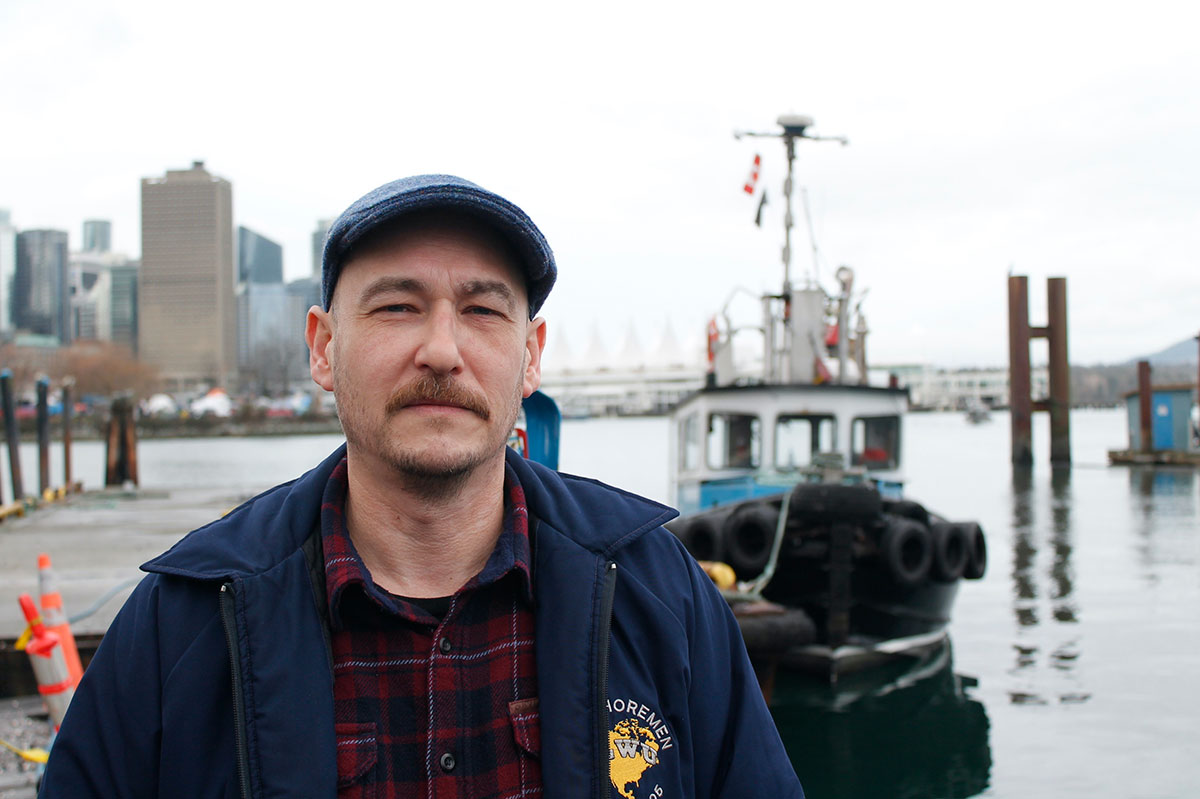 A man stands on a dock with a tugboat in the background. He has a serious expression and wears a cap and jacket.