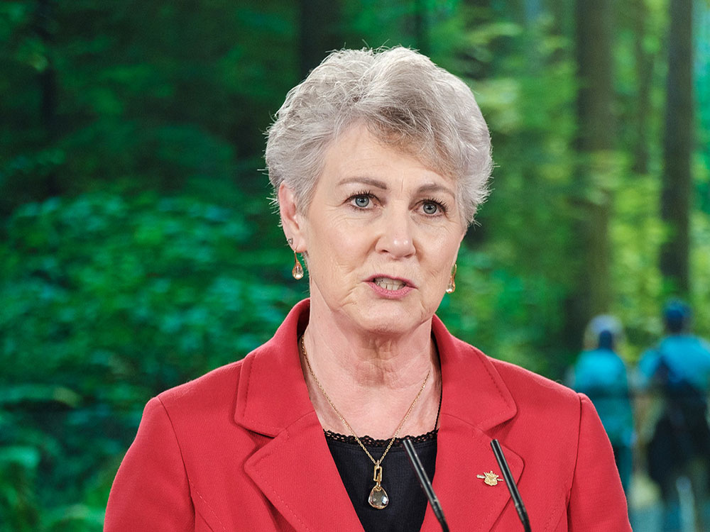 A woman with short silver hair is giving a speech in front of a forest background.
