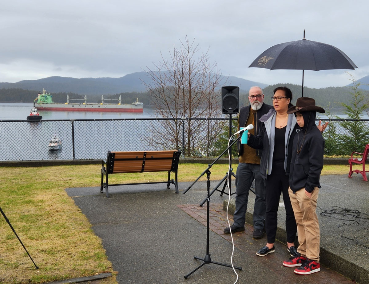 A woman wearing black stands at a microphone. On her left, is a man with a grey beard. On her right, a boy in is early teens wearing a bark hoodie and cowboy hat. It is grey and rainy and in the background is a freighter on the silver ocean.