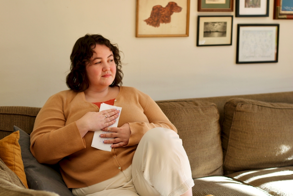 A woman sits on a couch, holding a handwritten letter to her chest. She looks off to her left.