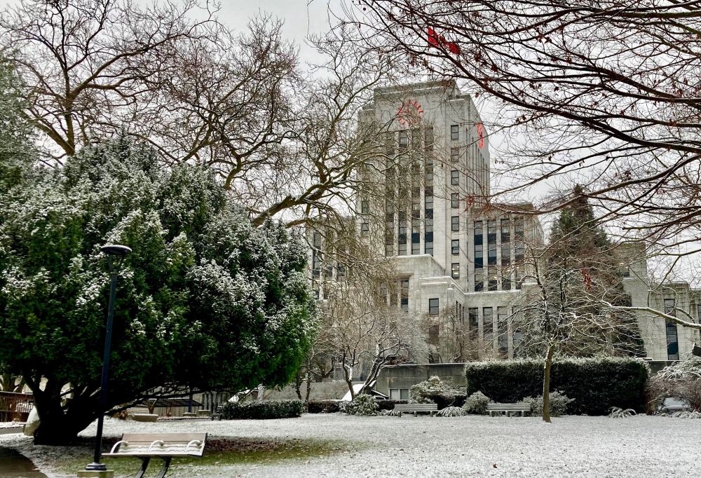 Vancouver’s City Hall building with trees and a bench in the foreground of the photo. A light dusting of snow covers the grounds and the trees.