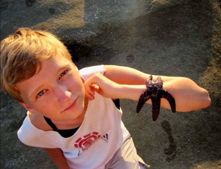 A family photo shows Dani Cooper on the beach. They are looking up at the camera, wearing a loose white T-shirt with the arms and neck cut away. There is a purple starfish on their forearm, which they are raising towards the camera.