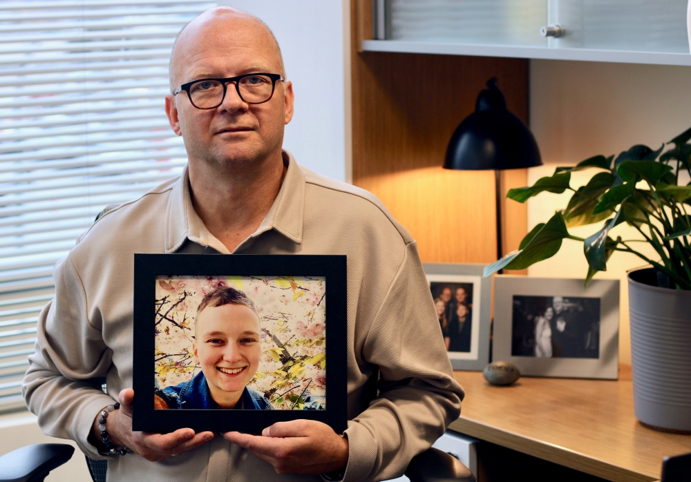 Dennis Cooper holds a framed portrait of their adult child, Dani Cooper. Dani has short blond hair and is smiling in front of a cherry blossom tree in the picture.