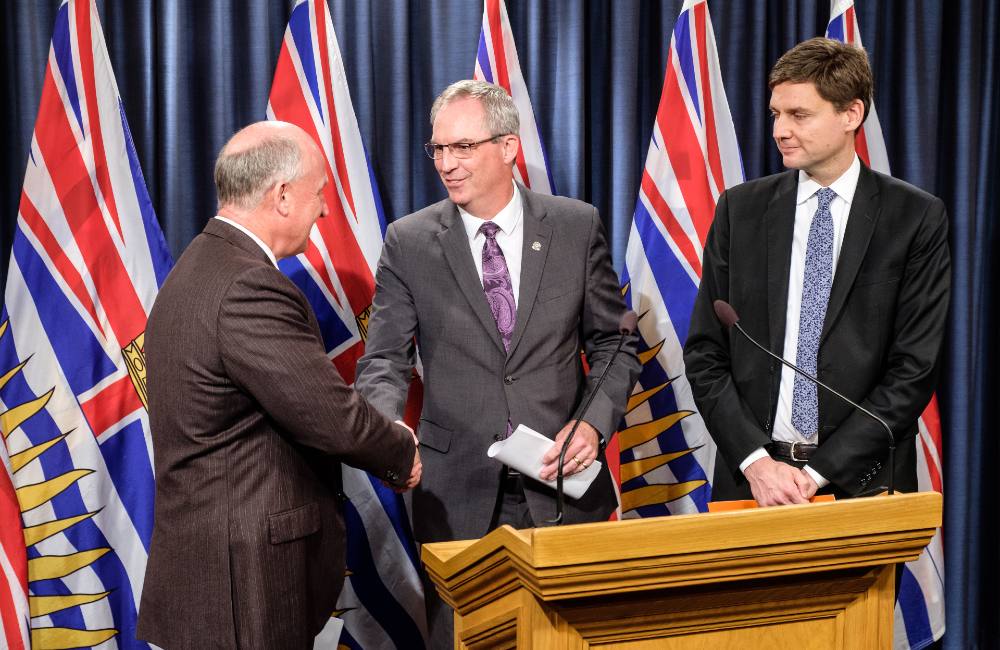 Three middle aged men in suits stand at a podium, two of the men shaking hands. Behind them are blue and red flags for the Province of B.C.