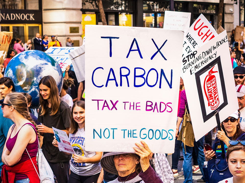 People march in a protest carrying signs that say “Tax carbon Tax the bads, not the goods” and “stop burning down the house.”