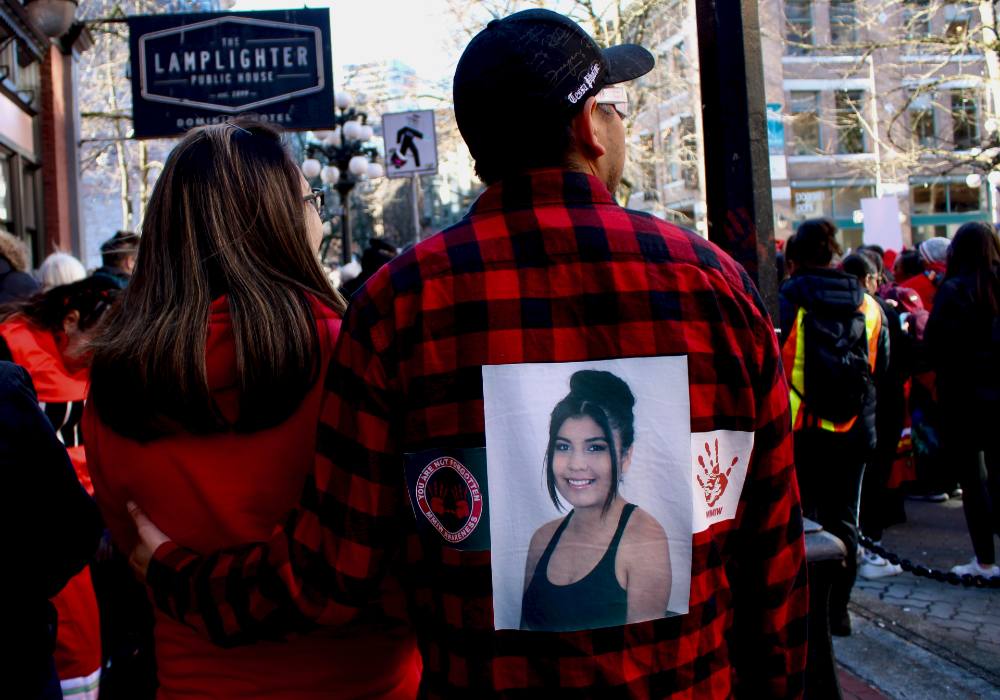 A man wearing a black ball cap and red-and-black check overshirt walks away from the camera, with his arm around a woman with long black hair. On his back is a large photo of a young woman.