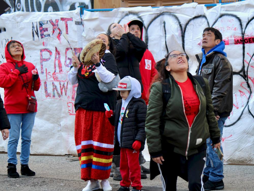 Seven Indigenous people, many dressed in red, are standing across the frame against the background of a white homemade parade banner. One person is holding a drum, a child is holding a frozen treat, and others are looking up at the sky in wonder at an eagle (out of frame) soaring overhead.  