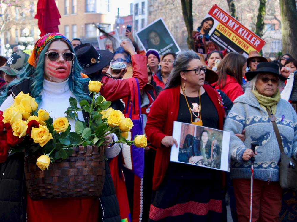 Indigenous women march toward the camera. One holds a basket of red and yellow roses. Another holds a photo of a young woman.